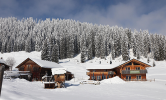 Eine malerische Winterlandschaft mit schneebedeckten Hütten. Hohe, grüne Nadelbäume umgeben die Szene unter einem klaren Himmel.