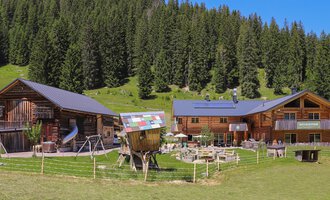 Ein malerisches Bergdorf mit traditionellen Holzhäusern und einer grünen Wiese. Umgeben von hohen Bäumen und klarem Himmel. | © Naturzeithof Café für dich | Stefanie Conticello