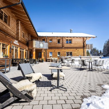 Ein gemütliches Holzgebäude im Winter mit einer Terrasse. Sessel und Tische stehen auf dem schneebedeckten Boden, umgeben von verschneiten Bäumen.