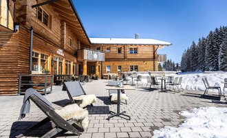 Ein gemütliches Holzgebäude im Winter mit einer Terrasse. Sessel und Tische stehen auf dem schneebedeckten Boden, umgeben von verschneiten Bäumen.