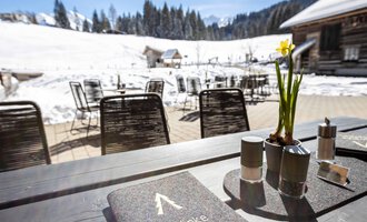A table outdoors with a menu and a small flower. In the background, snow-covered mountains and a cozy atmosphere can be seen.