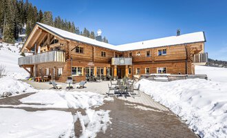 A cozy wooden house in the snow with a terrace and comfortable seating. In the background, there are snow-covered trees and a clear blue sky.
