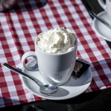 Eine Tasse mit Sahne auf einem weißen Unterteller. Der Hintergrund ist ein rot-weiß kariertes Tischläufer. | © Kleinwalsertal Tourismus | Dominik Berchtold