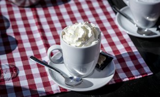 Eine Tasse mit Sahne auf einem weißen Unterteller. Der Hintergrund ist ein rot-weiß kariertes Tischläufer. | © Kleinwalsertal Tourismus | Dominik Berchtold