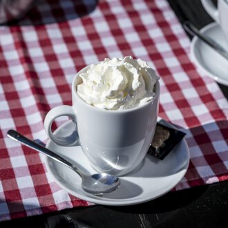 © Kleinwalsertal Tourismus | Dominik Berchtold A cup with cream and a spoon is on a table with a red-and-white checkered tablecloth. In the background, more cups can be seen. | © Kleinwalsertal Tourismus | Dominik Berchtold