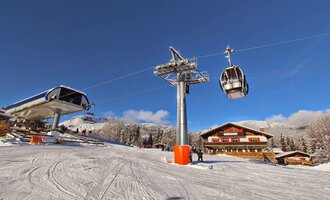 A ski lift station in a snowy mountain landscape. In the background, there is a chalet and a clear blue sky visible.