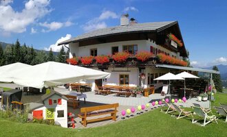 A cozy house in the mountains with colorful flower boxes and terraces with sun umbrellas. The surroundings are green and offer a beautiful view of the landscape. | © Cafe Walserblick | Marcus Dornach