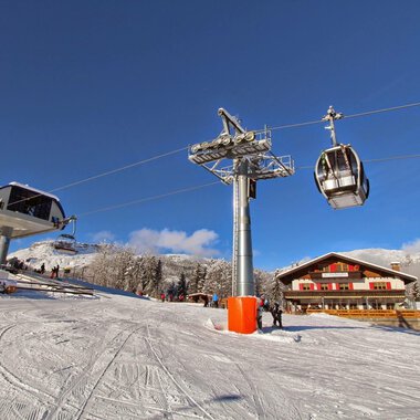 A ski lift station in a snowy mountain landscape. In the background, there is a chalet and a clear blue sky visible.