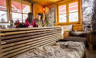 A cozy café with wooden furniture and soft blankets. Two people are sitting by the window and enjoying their time together. | © Kleinwalsertal Tourismus | Dominik Berchtold