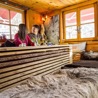 © Kleinwalsertal Tourismus | Dominik Berchtold Ein gemütliches Café mit Holzmöbeln und weichen Decken. Zwei Personen sitzen am Fenster und genießen ihre Zeit miteinander. | © Kleinwalsertal Tourismus | Dominik Berchtold