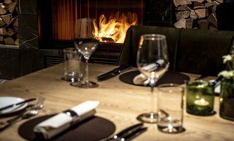Ein eleganter Tisch mit Geschirr und Gläsern, dekoriert für ein Abendessen. Im Hintergrund brennt ein Kamin, der eine gemütliche Atmosphäre schafft. | © A-ROSA Ifen Hotel Kleinwalsertal