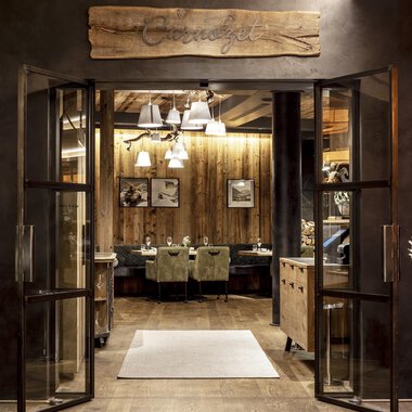 Ein einladender Eingang zu einem Restaurant mit Holzdetails und warmem Licht. Die Atmosphäre ist gemütlich und einladend. | © A-ROSA Ifen Hotel Kleinwalsertal