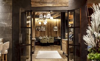 Ein einladender Eingang zu einem Restaurant mit Holzdetails und warmem Licht. Die Atmosphäre ist gemütlich und einladend. | © A-ROSA Ifen Hotel Kleinwalsertal