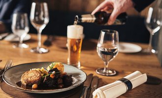 Ein gedeckter Tisch mit einem Teller köstlichem Essen und einem Glas Bier. Eine elegante Atmosphäre mit Wein- und Wassergläsern. | © A-ROSA Ifen Hotel Kleinwalsertal