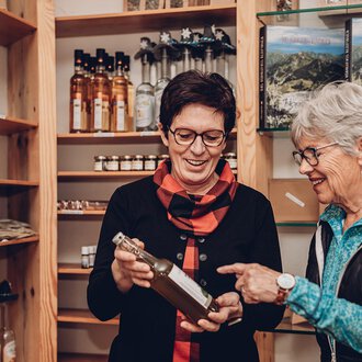 Zwei Frauen betrachten gemeinsam eine Flasche in einem Laden. Im Hintergrund sind Regale mit weiteren Produkten zu sehen. | © Carolin Ries | www.calejofotografie.de