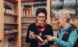 Zwei Frauen betrachten gemeinsam eine Flasche in einem Laden. Im Hintergrund sind Regale mit weiteren Produkten zu sehen. | © Carolin Ries | www.calejofotografie.de