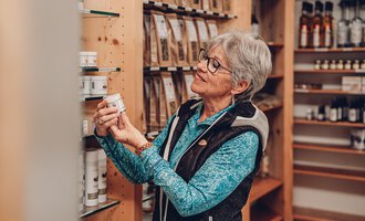Eine ältere Frau schaut sich ein Produkt in einem Geschäft an. Der Raum ist gut beleuchtet und voller natürlicher Produkte. | © Carolin Ries | www.calejofotografie.de