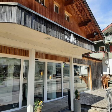 A modern building with wooden cladding stands in the foreground. There are large windows and a relaxed atmosphere in the surroundings. | © das Wallis Restaurant & Brasserie | Veronika Senn