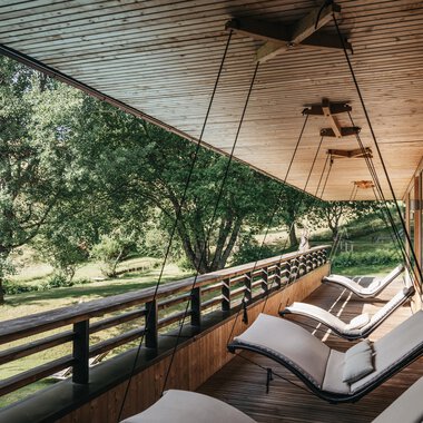 A relaxing veranda with modern loungers overlooking a green garden. The wooden ceiling and the natural surroundings create a peaceful atmosphere. | © Naturhotel Chesa Valisa | Heldentheater