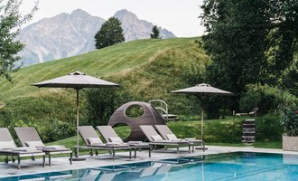 Ein Poolbereich mit Liegen und Sonnenschirmen. Im Hintergrund sind sanfte Hügel und Berge zu sehen.