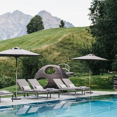 Ein Poolbereich mit Liegen und Sonnenschirmen. Im Hintergrund sind sanfte Hügel und Berge zu sehen.
