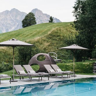 A pool area with loungers and sun umbrellas. In the background, gentle hills and mountains can be seen.