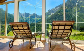 Two lounge chairs are positioned in front of large windows overlooking green mountains. The room feels inviting and bright. | © Hotel Leitner