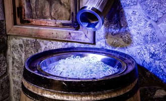 A wooden barrel with ice cubes stands in a cozy environment. In the background, there are stone walls and a wooden window frame visible. | © Hotel Leitner