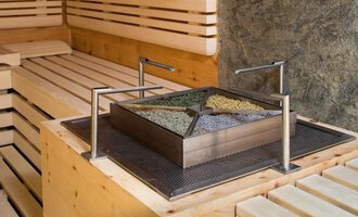 A modern sauna area with a square container that contains various herbs. The walls and benches are made of light wood. | © Hotel Leitner