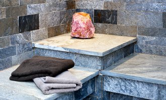 A modern sauna interior with gray stone walls. On one of the stones lies a pink salt block and some terry towels. | © Hotel Leitner