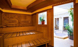 A cozy wooden sauna with a bench and large windows. Through the door, you can see a small indoor pool and plants in the background. | © Hotel Leitner