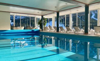 Ein heller Innenpool mit klarem Wasser und Liegen daneben. Große Fenster lassen viel Tageslicht herein und bieten einen Blick nach draußen. | © Hotel Leitner