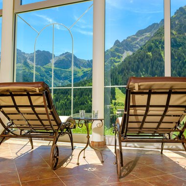 Zwei Liegestühle stehen vor großen Fenstern mit Blick auf grüne Berge. Der Raum wirkt einladend und hell. | © Hotel Leitner