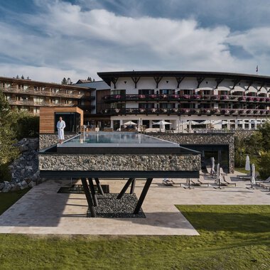 A modern hotel with a large glass pool is in the foreground. The surrounding landscape is green and inviting. | © A-ROSA Ifen Hotel Kleinwalsertal