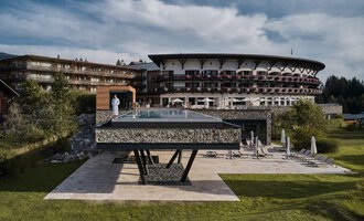 A modern hotel with a large glass pool is in the foreground. The surrounding landscape is green and inviting. | © A-ROSA Ifen Hotel Kleinwalsertal