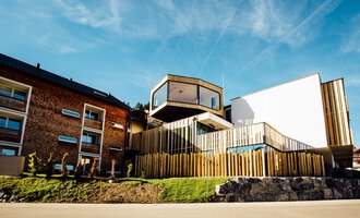Ein modernes Gebäude mit Holzverkleidung und klaren Linien. Es befindet sich in einer grünen Umgebung, umgeben von anderen Häusern. | © Genuss- & Aktivhotel Sonnenburg |Heike Wohlgenannt