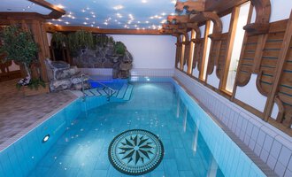 An elegant indoor pool with clear blue water and decorative tiles. Wooden ornaments and plants adorn the wall, creating a tranquil atmosphere. | © Hotel Alpenstüble Appartements | Manuel Riezler