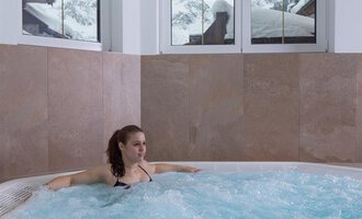 A woman relaxes in a hot tub. In the background, there are windows with a view of snowy mountains. | © Hotel Alpenstüble Appartements | Manuel Riezler