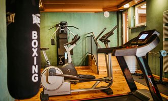A modern gym with various training equipment. In the foreground, a treadmill and a punching bag are visible. | © Hotel Alpenstüble KG