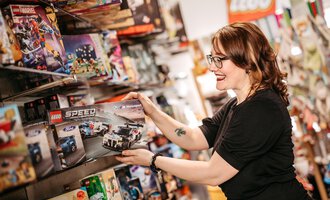 Eine Frau hält ein LEGO-Set der Speed-Serie in einem Spielzeugladen. Sie lächelt, während sie die Regale mit verschiedenen LEGO-Sets betrachtet. | © Sascha Gast, Dein Fotograf