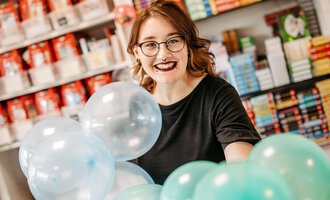 Eine junge Frau mit Brille lächelnd, während sie bunte Luftballons hält. Im Hintergrund sind Regale mit Büchern sichtbar. | © Sascha Gast