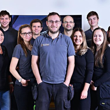 Eine Gruppe von zwölf Personen steht zusammen und lächelt. Sie tragen ähnliche graue Arbeitskleidung und befinden sich in einem hellen Raum. | © Der Elektriker Markus Leitgeb GmbH