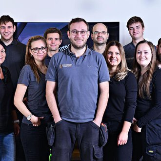 © Der Elektriker Markus Leitgeb GmbH A group of twelve people stands together and smiles. They are wearing similar gray work clothing and are in a bright room. | © Der Elektriker Markus Leitgeb GmbH