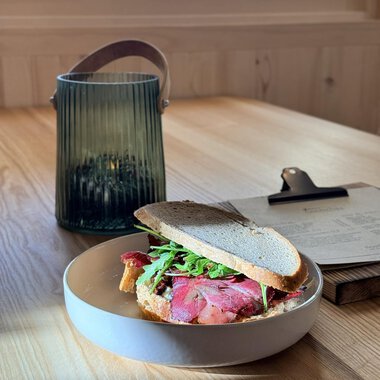 A sandwich with roast beef and arugula on a plate. In the background, there is a green lantern on a wooden table. | © Einkehr Guntschau