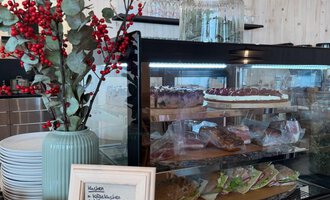 An inviting café area with a glass display case full of pastries and snacks. Next to the display case stands a vase with red berries. | © Einkehr Guntschau