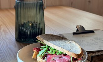 Ein Sandwich mit Rinderbraten und Rucola auf einem Teller. Im Hintergrund steht eine grüne Laterne auf einem Holztisch. | © Einkehr Guntschau