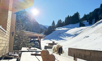 Eine ruhige Winterlandschaft mit Schnee und Bergen im Hintergrund. Auf einer Holzterrasse stehen einige Werkzeuge und es scheint die Sonne. | © Einkehr Guntschau