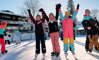 A group of cheerful children stands on an ice rink, raising their hands in the air. They are wearing colorful winter clothing and enjoying the sunny winter day. | © Kleinwalsertal Tourismus | Oliver Farys