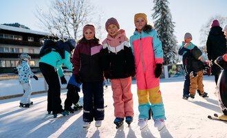 Drei Mädchen stehen auf einer Eisbahn, umgeben von weiteren Kindern, die Schlittschuhfahren. Es ist ein sonniger Tag mit klarem Himmel und Schnee in der Umgebung. | © Kleinwalsertal Tourismus | Oliver Farys