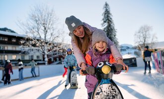 Eine Mutter und ihr Kind fahren auf einer Pinguin-Eisfläche. Im Hintergrund sind winterliche Landschaften und Menschen beim Eislaufen zu sehen. | © Kleinwalsertal Tourismus | Oliver Farys
